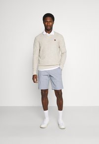 Beige knitted sweater with round neck, gray patterned shorts, and white sneakers; model stands against a plain background.