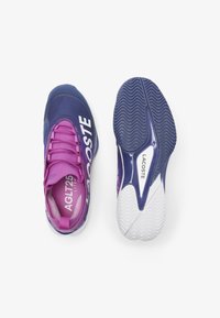 Scarpe da atletica viola e rosa con una superficie testurizzata, caratterizzate dal logo "LACOSTE" e da una suola in gomma bianca con un motivo a zigzag.