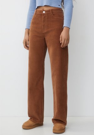 Person wearing high-waist brown corduroy pants, tan shoes, and a cropped light blue ribbed sweater, standing against a plain white background.