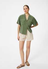 Grünes Kurzarm-Hemd mit Knopfleiste und zwei Brusttaschen, kombiniert mit beigen Shorts und gestreiften Sandalen. Schlichte, lockere Passform und strukturierter Stoff.