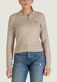 Beige kabelgebreide trui met kraag, drie knopen en een rood hartaccent op de borst. Getailleerd ontwerp met geribbelde zoom en manchetten.