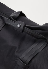 Svart duffelväska med textilstruktur, som har släta läderremmar, knappstängning och en stilren, strukturerad design.