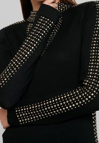 Top negro de manga larga con cuello alto, adornado con tachuelas doradas dispuestas en filas en los hombros y las mangas. Textura de punto suave.