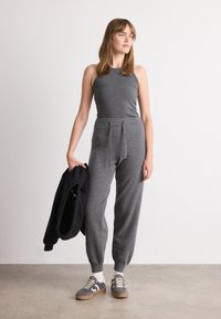 Grå ribbet tanktop kombinert med grå taperede joggebukser med knytebelte. Modellen holder en svart jakke og har på seg grå sneakers.
