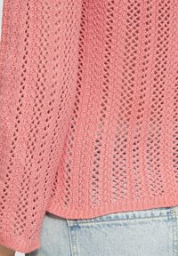 Rosa gestrickter Pullover mit einem offenen Strickmuster, locker geschnitten und mit gerippten Bündchen, kombiniert mit hellblauer Jeans.