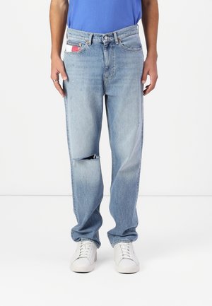 Jean bleu clair droit avec un genou gauche déchiré, porté avec des baskets blanches et une chemise bleue rentrée.