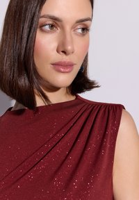 Bordowa sukienka o drapowanym kroju z błyszczącymi detalami, z plisami na ramieniu i gładką fakturą tkaniny.