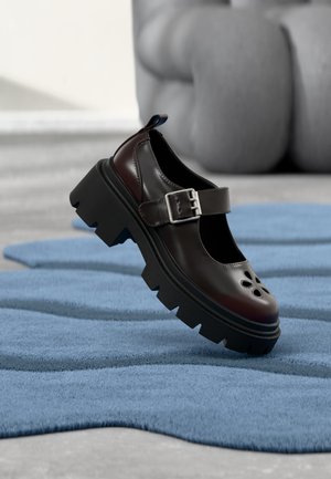 Black Mary Jane shoe with chunky sole, silver buckle strap, and floral cutouts, displayed on a textured blue surface indoors.