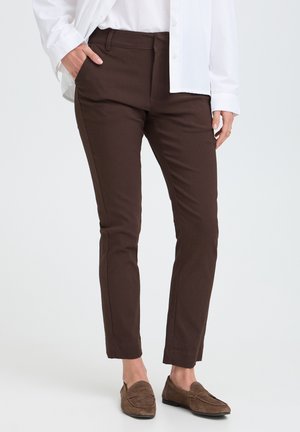 Personne portant un pantalon ajusté marron foncé, des mocassins marron clair et une chemise blanche à manches longues, une main dans la poche.