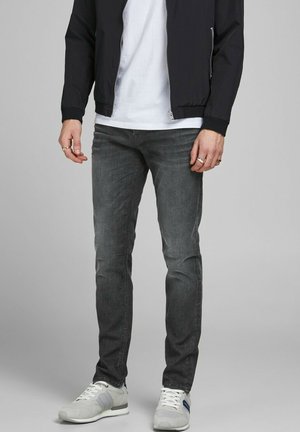 Mężczyzna ubrany w wyblakłe czarne obcisłe dżinsy, białe trampki, białą koszulkę i czarną kurtkę na zamek na jednolitym tle.