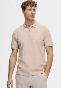 Polo shirt rosa chiaro con maniche corte, un colletto a due bottoni e cuciture bianche sottili lungo il colletto. La trama del tessuto è morbida e visibile.