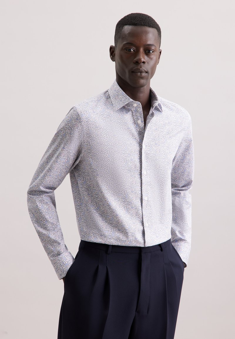 Camicia a maniche lunghe con bottoni in colori chiari con un motivo floreale, realizzata in un tessuto morbido. Abbinata a pantaloni scuri a pieghe.