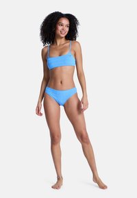 Σετ μπικίνι γαλάζιο με ανοιχτόχρωμο τοπ με λεπτές τιράντες και matching σλιπ με χαμηλή μέση, διακοσμημένο με σχέδια κυμάτων.