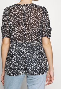 Blouse noire à fleurs avec manches courtes bouffantes, en tissu texturé, ornée de délicates fleurs blanches et lavande, avec un détail dos en forme de goutte.