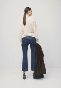Vrouw met een lange, donkere paardenstaart, gekleed in een crème coltrui, blauwe cropped jeans en zwarte hakken, die een bruine bontjas vasthoudt tegen een effen lichte achtergrond.