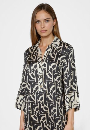 Woman with straight brown hair wearing a black and white abstract patterned button-up blouse with three-quarter sleeves.