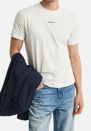 Jasna kremowa koszulka bawełniana z okrągłym dekoltem i krótkimi rękawami, z niebieskim logo "GANT". W połączeniu z niebieskimi dżinsami i granatowym żakietem.