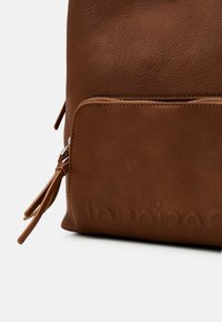 Mochila de cuero marrón con una superficie texturizada, un bolsillo frontal con cremallera y un detalle decorativo de borlas en la cremallera. Marca sutilmente grabada.