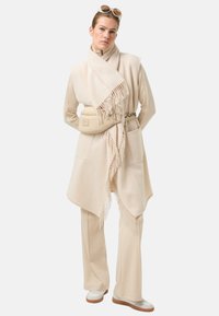 Béžový pléd s třásněmi na okrajích, doplněný béžovými kalhotami a krémovými botami. Součástí jsou také ledvinka a velké sluneční brýle pro zvýraznění detailů.