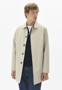 Bež plašč iz lahkega materiala z ovratnikom, sprednjimi gumbi in stranskimi žepi. Obut nad temnim puloverjem in kavbojkami.