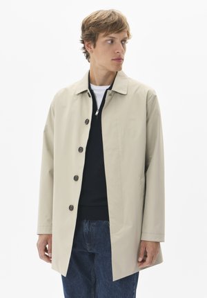 Világos bézs színű trench coat könnyű anyagból, gallérral, elől gombokkal és oldalsó zsebekkel. Sötét pulóver és farmernadrág felett viselve.