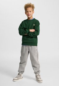 Grön sweatshirt med ribbade muddar, kombinerad med ljusgrå koniska sweatpants, båda plaggen prydda med en logotyp och kompletterade med beige sneakers.