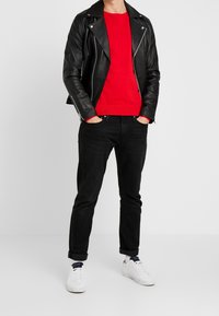 Veste de motard en cuir noir portée sur un pull rouge, associée à un jean noir et des baskets blanches. La veste est dotée de ferrures et de fermetures éclair argentées.