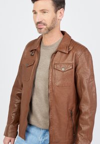 Braune Lederjacke mit Hemdkragen, zwei vorderen Taschen und einer seitlichen Reißverschlusstasche. Glatte Oberfläche mit Nahtdetails.