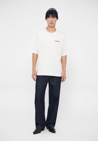 Camiseta blanca de algodón con mangas cortas y texto de logo negro. Combinada con pantalones vaqueros de pierna ancha de color azul oscuro y un gorro de patrón.
