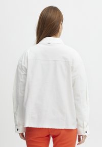 Hvid denimjakke med en afslappet pasform, synlige syninger og knaplukning ved ærmerne. Kantene er skåret ujævnt for en lagdelt effekt.