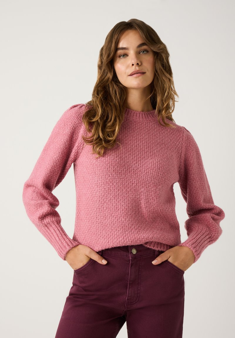 Rosa gestrickter Pullover mit strukturiertem Muster, rundem Halsausschnitt und gerippten Bündchen, kombiniert mit dunkelvioletten hochgeschnittenen Jeans.