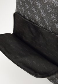 Sac à main noir et gris avec un extérieur à motifs, doté d'une fermeture éclair. L'intérieur est tapissé de noir et présente un motif de logo "GUESS" répété.