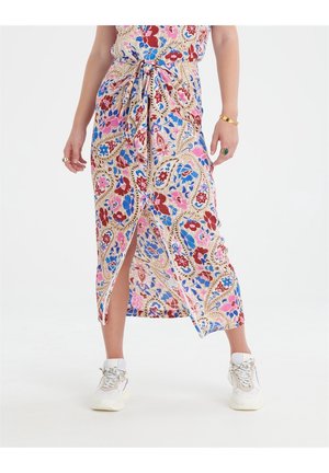 Kvinde iført en blomsterprintet maxinederdel med frontslids, hvide chunky sneakers, guldt armbånd og ringe på hænderne.