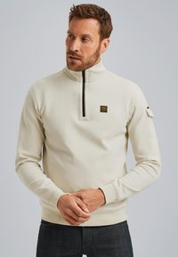 Sudadera de color crema con un cuello alto y cremallera negra, que presenta un pequeño parche con logo en el pecho y una tela texturizada.