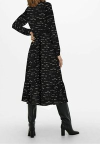 Černé dlouhé šaty s bílým vzorem textu, volný střih a vrstvený spodní lem. Kombinováno s černými botami do kolen.