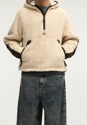 Personne portant un sweat à capuche polaire beige avec une poche avant et des manches bordées de foncé, associé à un jean bleu ample sur fond uni.