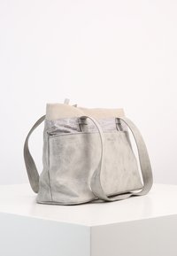 Borsa a tracolla in pelle texturizzata grigia chiara con manici lunghi, dettagli in tessuto argentato e una morbida parte superiore beige, appoggiata su un piedistallo bianco.