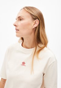 Beige katoenen T-shirt met een ronde hals, korte mouwen en een rood geborduurd bloemenlogo op de borst. Gladde textuur.
