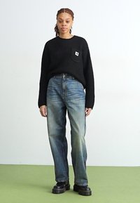 Maglione nero a coste con tasca, abbinato a jeans dritti blu sbiaditi e scarpe con plateau nere su un pavimento verde chiaro.