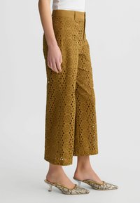Světle hnědé kalhoty culottes vyrobené z perforované tkaniny, s kruhovými výřezy, vysokým pasem a širokými nohavicemi, doplněné o vzorované boty.