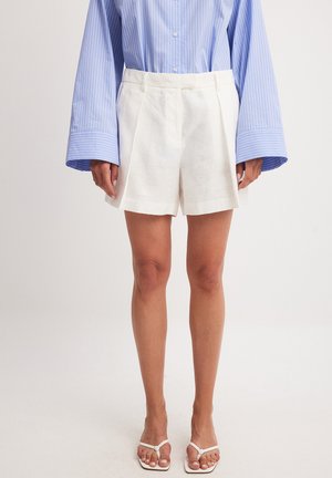 Weisse Leinen-Shorts mit Faltenentwurf, kombiniert mit einem oversized Hemd in Blau und Weiss gestreift, das eine lässige Passform und Struktur zeigt.
