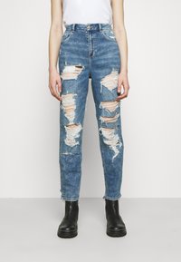 Sprängda blå denimjeans med hög midja, med flera slitna lappar och fransade kanter, tillsammans med svarta ankelstövlar.