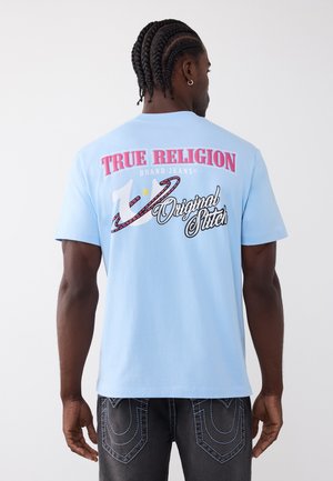 Man met gevlochten haar draagt een lichtblauw True Religion t-shirt en donkere spijkerbroek, met de rug naar de kijker, tegen een effen lichte achtergrond.