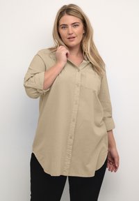 Chemise beige à manches longues, avec une poche poitrine et un ourlet arrondi, fabriquée dans un tissu léger au toucher doux.