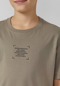 Oliwkowozielona bawełniana koszulka z okrągłym dekoltem i krótkimi rękawami. Czarny nadruk tekstowy w formie prostokąta z hasłami związanymi z podróżami.