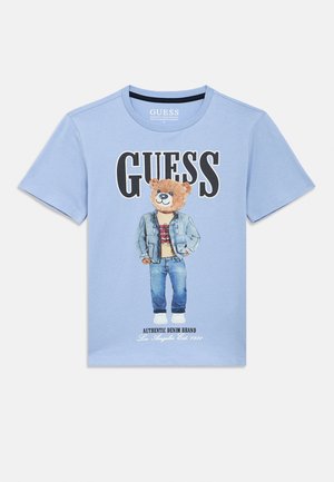 Tricou din bumbac albastru deschis, cu un desen cu urs purtând o jachetă din denim și blugi. Text îndrăzneț "GUESS" deasupra și detalii ale brandului dedesubt, în negru.