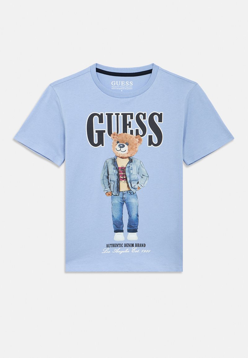 Camiseta de algodón azul claro con un gráfico de un oso con chaqueta vaquera y vaqueros. Texto destacado «GUESS» en la parte superior y detalles de la marca en negro debajo.