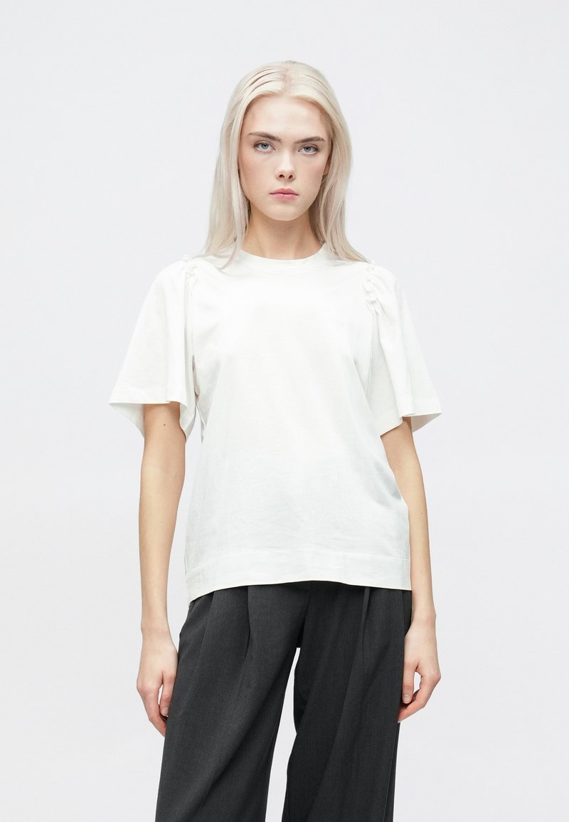 Hvit T-skjorte med korte, løse ermer og rynkedetaljer på skuldrene. Stoffet er glatt med en liten glans, kombinert med grå bukser.