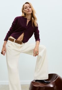 Femme portant un cardigan violet, un pantalon large blanc, une ceinture marron et des bottes noires, reposant un pied sur une chaise marron brillante.