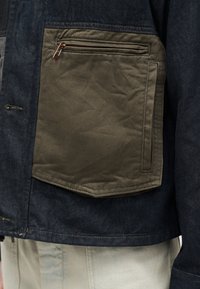 Σκούρο denim μπουφάν με μια μεγάλη τσέπη με φερμουάρ σε ελιά πράσινο χρώμα στο πλάι και ανοιχτό μπεζ παντελόνι που φαίνεται από κάτω από το μπουφάν.